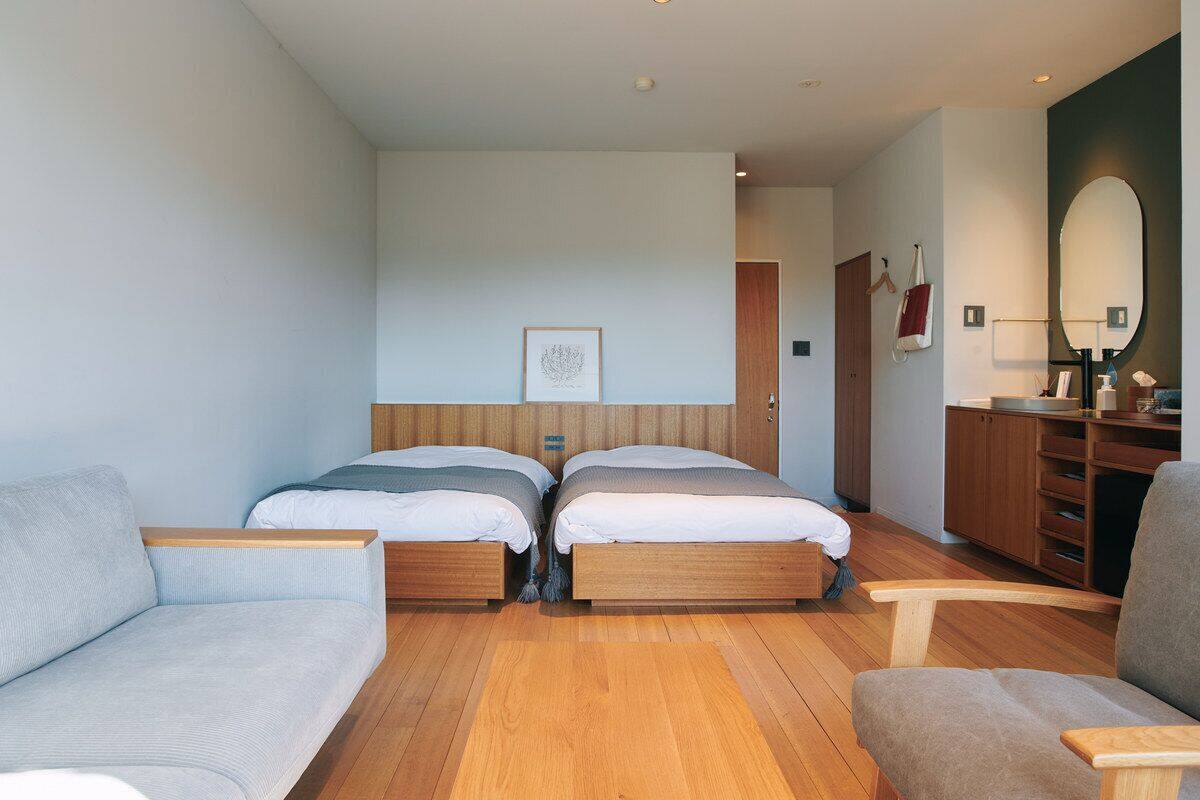 A modern hotel room with two single beds, a sofa, a chair, a wooden dresser, and a large mirror.