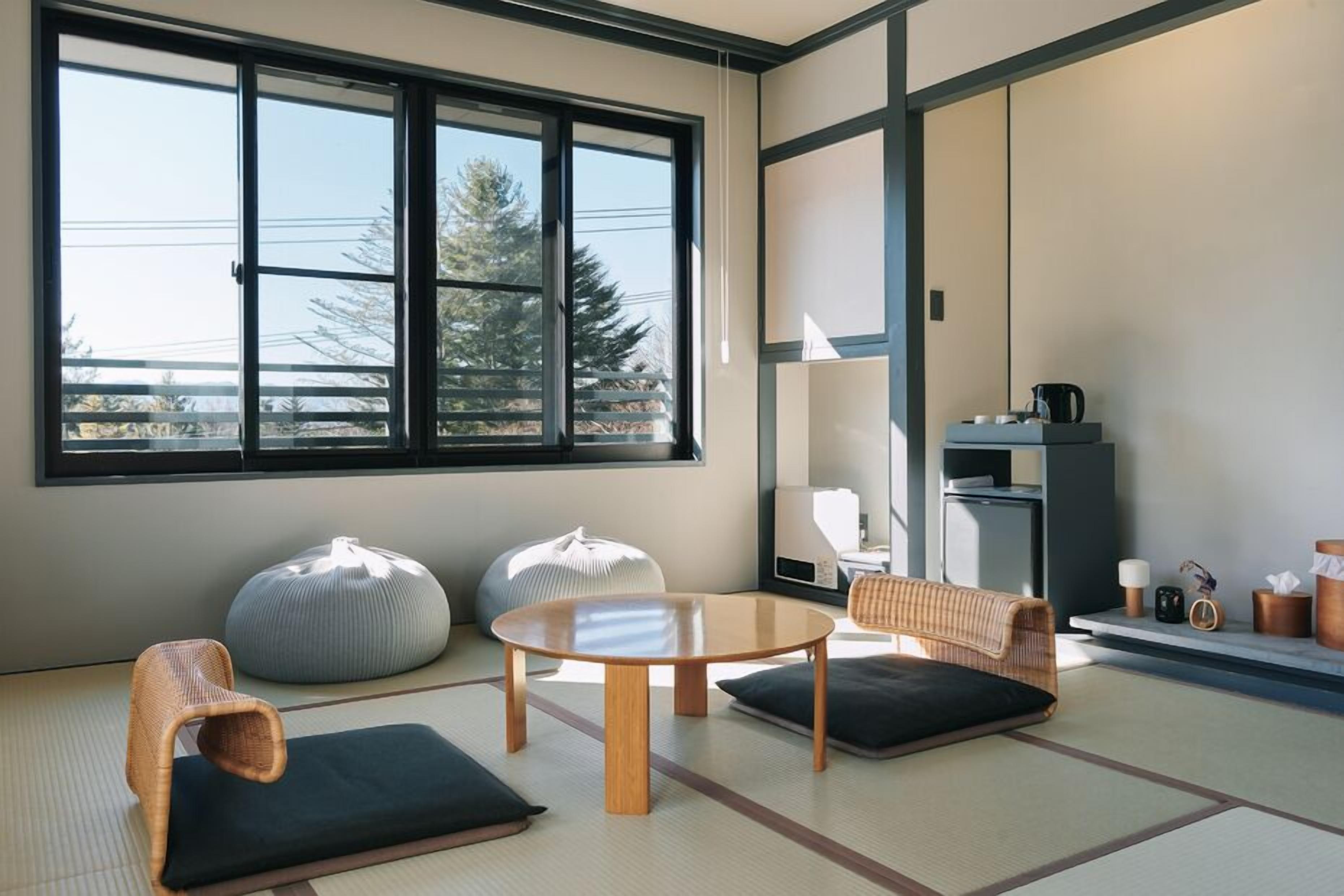 Una habitación con suelo de tatami, una mesa redonda, dos cojines, una silla de fibras naturales y un ventanal con vistas a árboles.