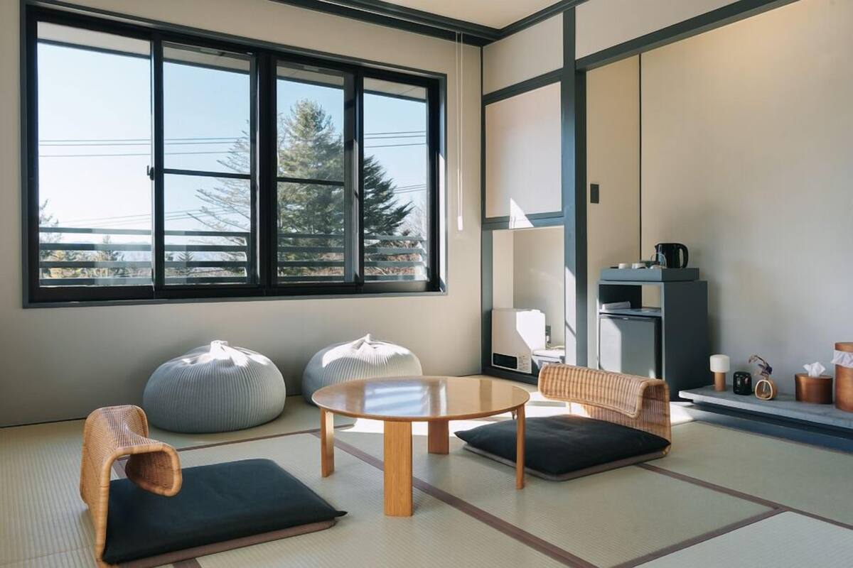 A room with tatami flooring, a round table, two cushions, a woven chair, and a large window with a view of trees.