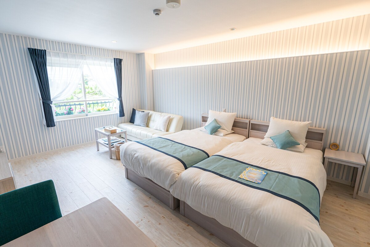 A double bed with a blue accent, a small bedside table, a window with curtains, and a view of greenery outside.