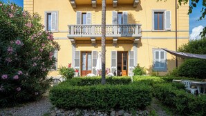 Ein Gebäude mit gelber Fassade, mehreren Fenstern mit Fensterläden, einem Balkon und einem Gartenbereich mit Tisch und Stühlen.