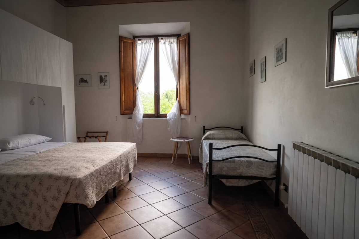 A bedroom with two beds, a window with curtains, a radiator, and a small stool.