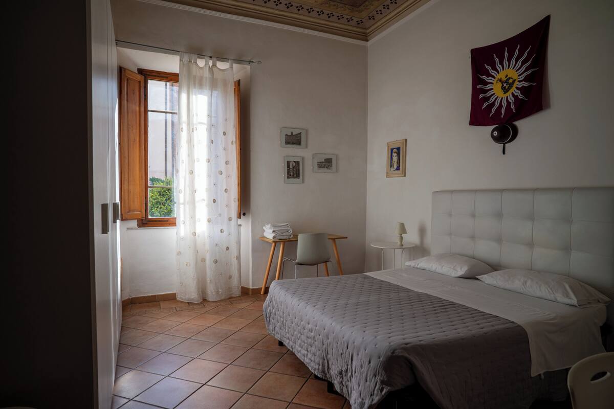 A bedroom with a bed, a window with curtains, a small table with a chair, and a wall-mounted lamp.
