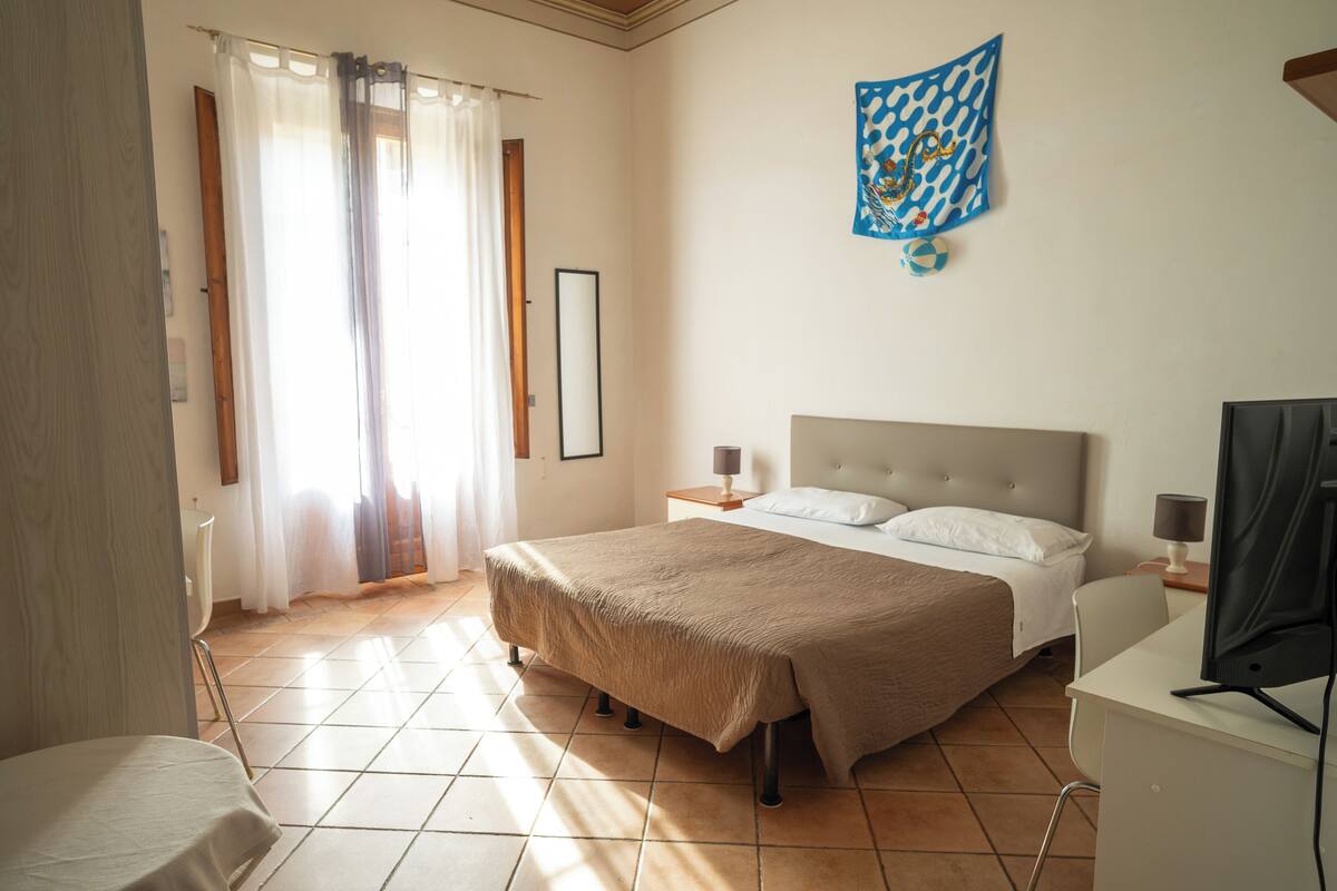 A hotel room with a bed, a desk, a chair, a television, a nightstand, a lamp, a window with curtains, and a wall decoration.