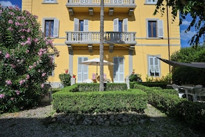 Ein gelbes Gebäude mit Balkonen, einer Steinmauer und einem Garten mit rosa Blumen.