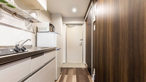 A compact kitchen with a built-in refrigerator, a sink with a faucet, and a microwave. The kitchen has a wooden floor and a door leading to another room. - Mr. Kinjo In Maehara (Ginowan)