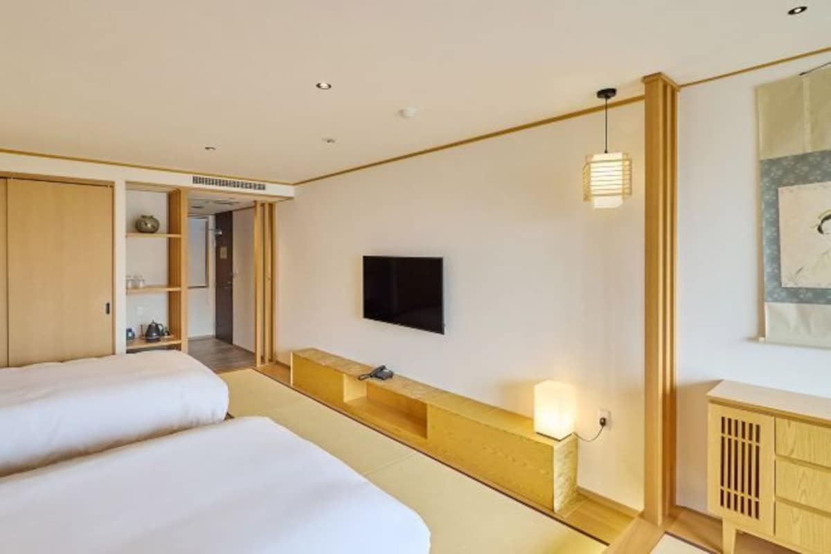 Habitación de hotel con dos camas, un televisor montado en la pared, una mesita de noche con lámpara y un armario de madera.