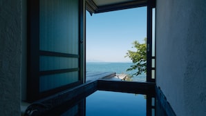 Blick aus einem Zimmer mit Holzboden und Fenster auf ein Gewässer, einen Steg und entfernte Berge.