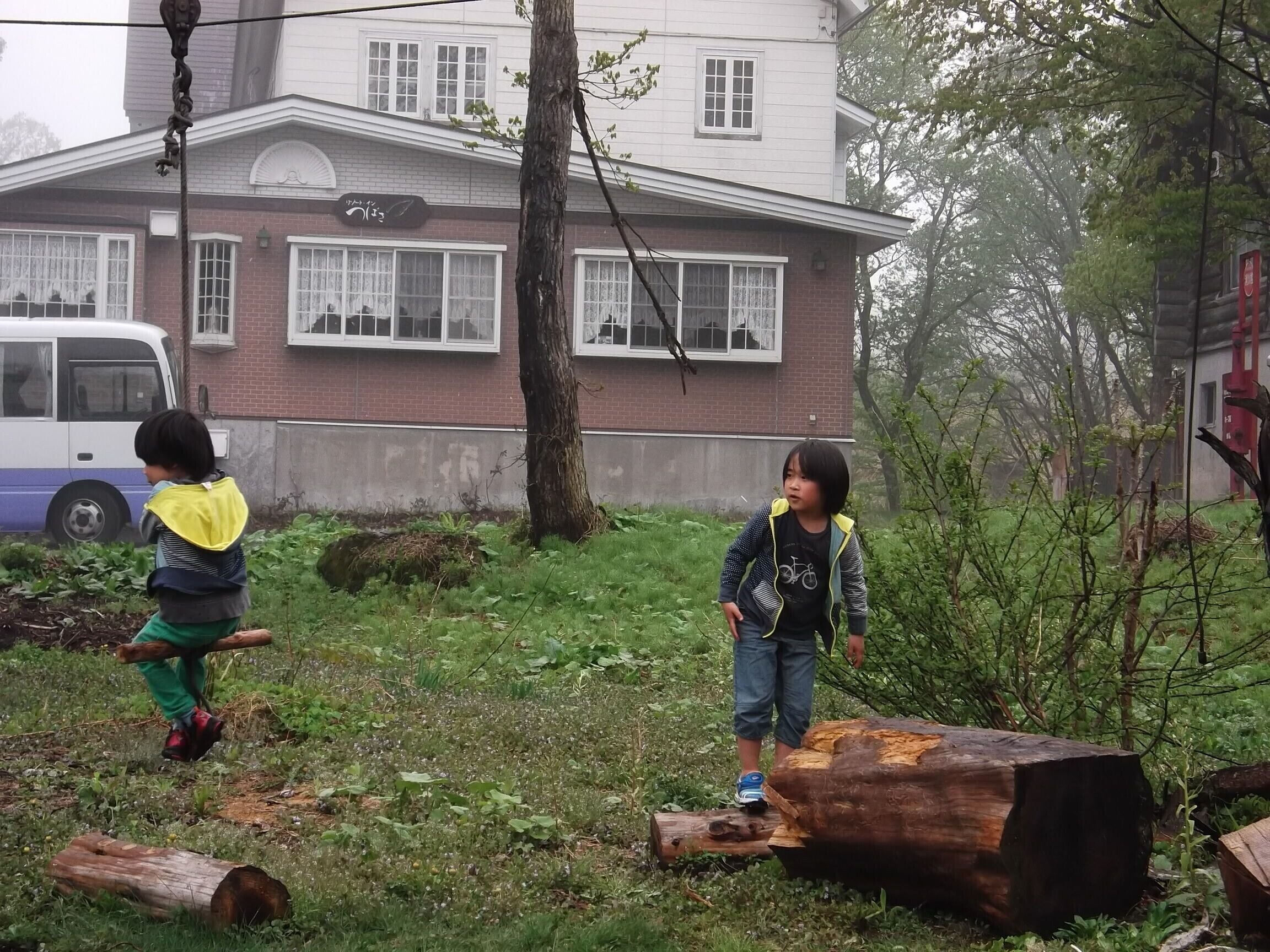 Two children playing in a grassy area with a large log and a building in the background.