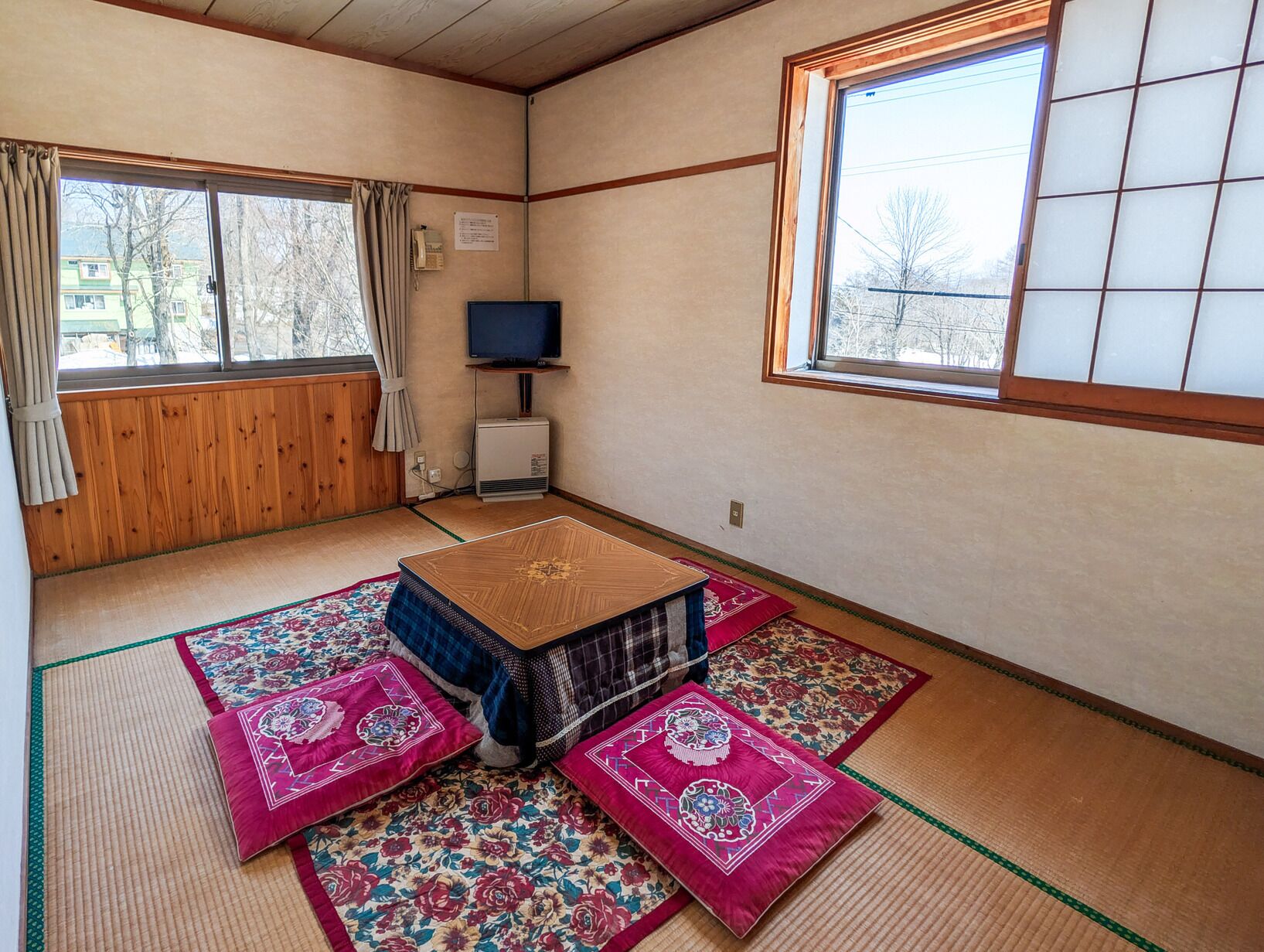 A traditional Japanese room with tatami flooring, a tatami mat table, a television, and a window with a view of trees.