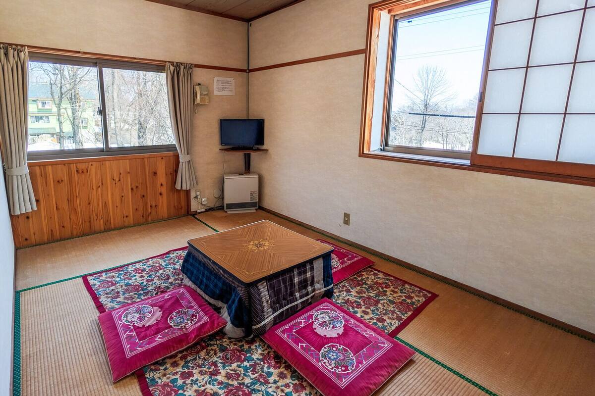 Une pièce de style japonais traditionnel, avec un sol recouvert de tatamis, une table basse en tatami, un téléviseur et une fenêtre donnant sur des arbres.