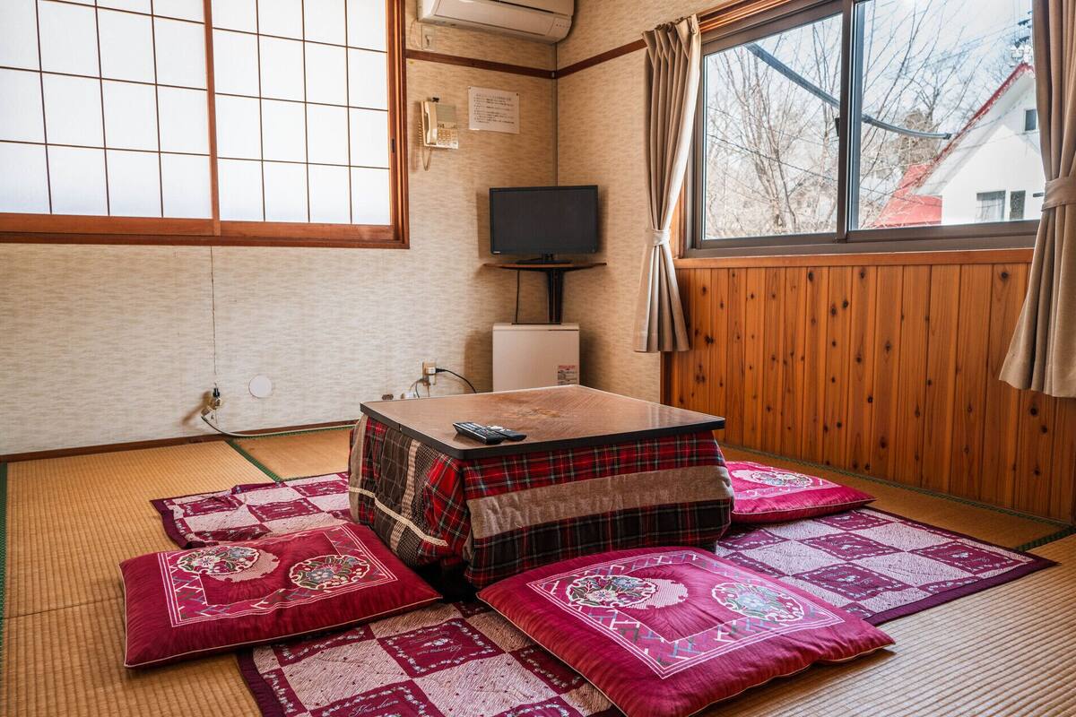Une pièce de style japonais traditionnel, avec un sol recouvert de tatamis, une basse table en bois entourée de coussins, un téléviseur et une fenêtre donnant sur un bâtiment à l’extérieur.