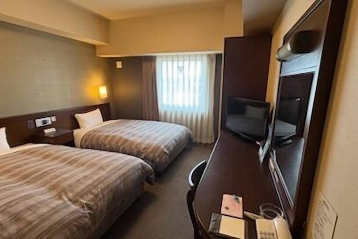Ein Hotelzimmer mit zwei Betten, einem Schreibtisch, einem Fernseher und einem Fenster mit Vorhängen.