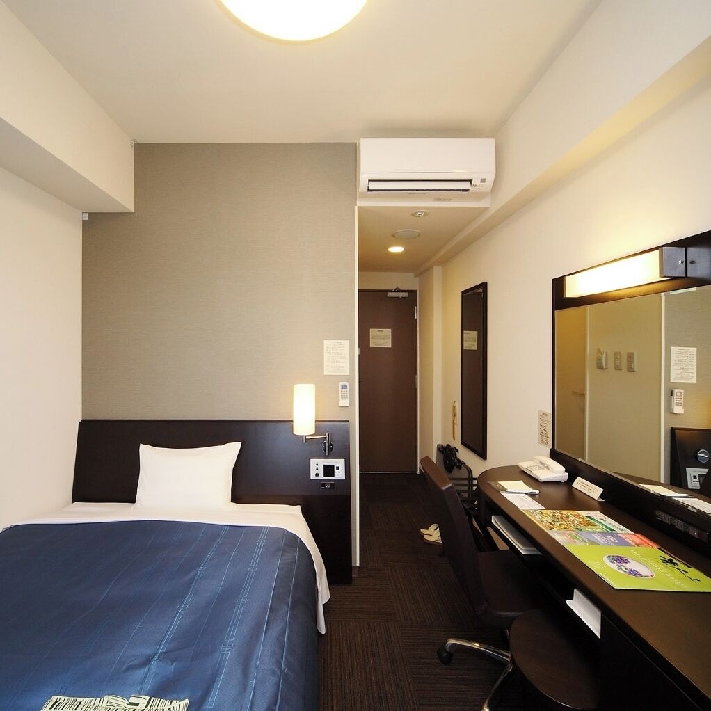 A hotel room with a bed, a desk, a mirror, an air conditioning unit, and a door leading to another room.