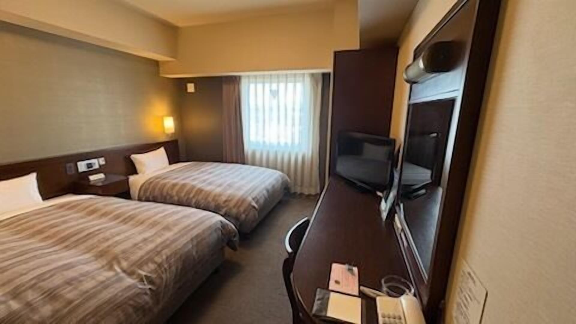 A hotel room with two beds, a desk with a chair, a television, and a window with curtains.