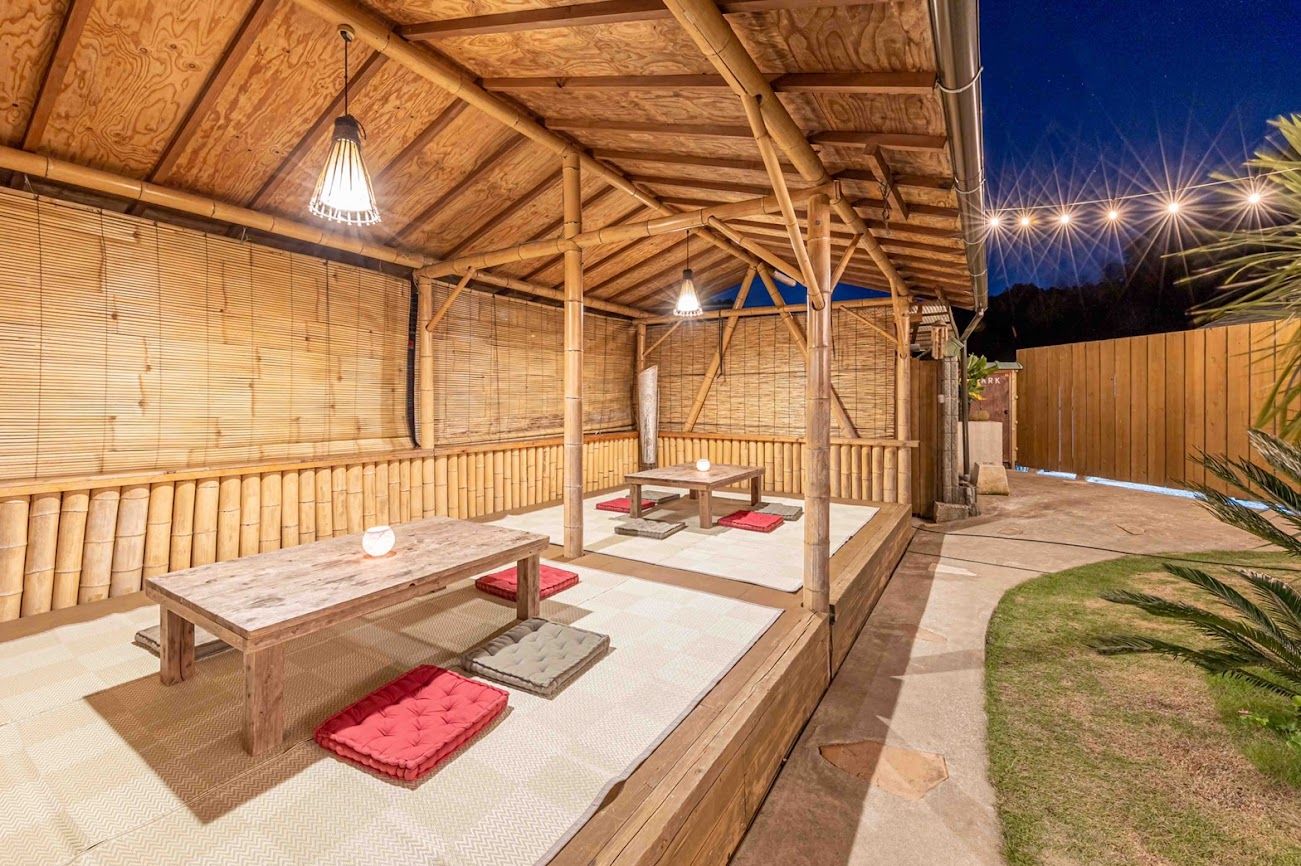 An outdoor covered patio with a wooden table and chairs, bamboo walls, and string lights.