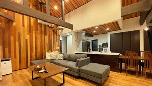 A modern living room with a sectional sofa, a wooden coffee table, and a kitchen area with dark cabinets and a countertop. - Kyoran Fukatei (Tateyama)