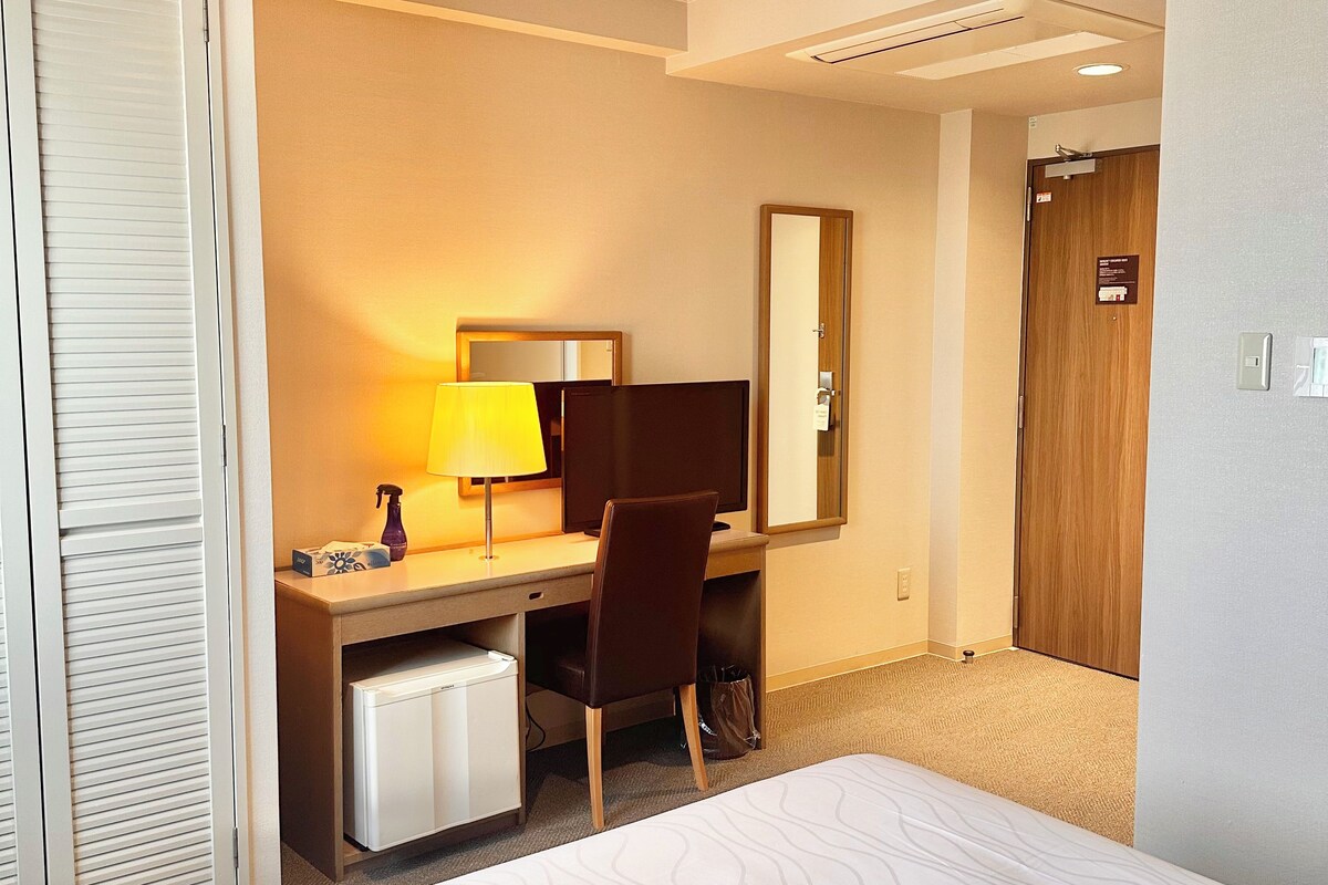 Quarto de hotel com cama, escrivaninha, cadeira, luminária de mesa, espelho, guarda-roupa e porta.