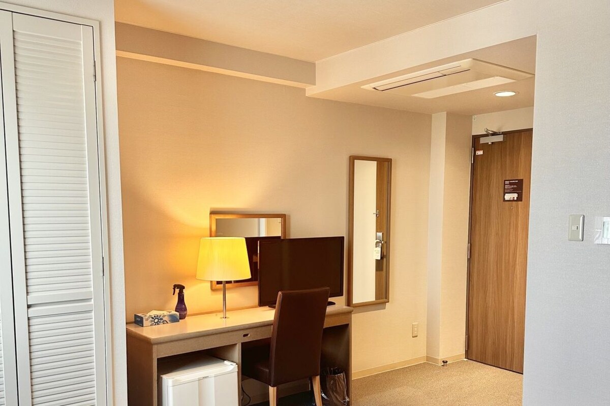 Quarto de hotel com cama, secretária com computador, cadeira, candeeiro, espelho e guarda-roupa.