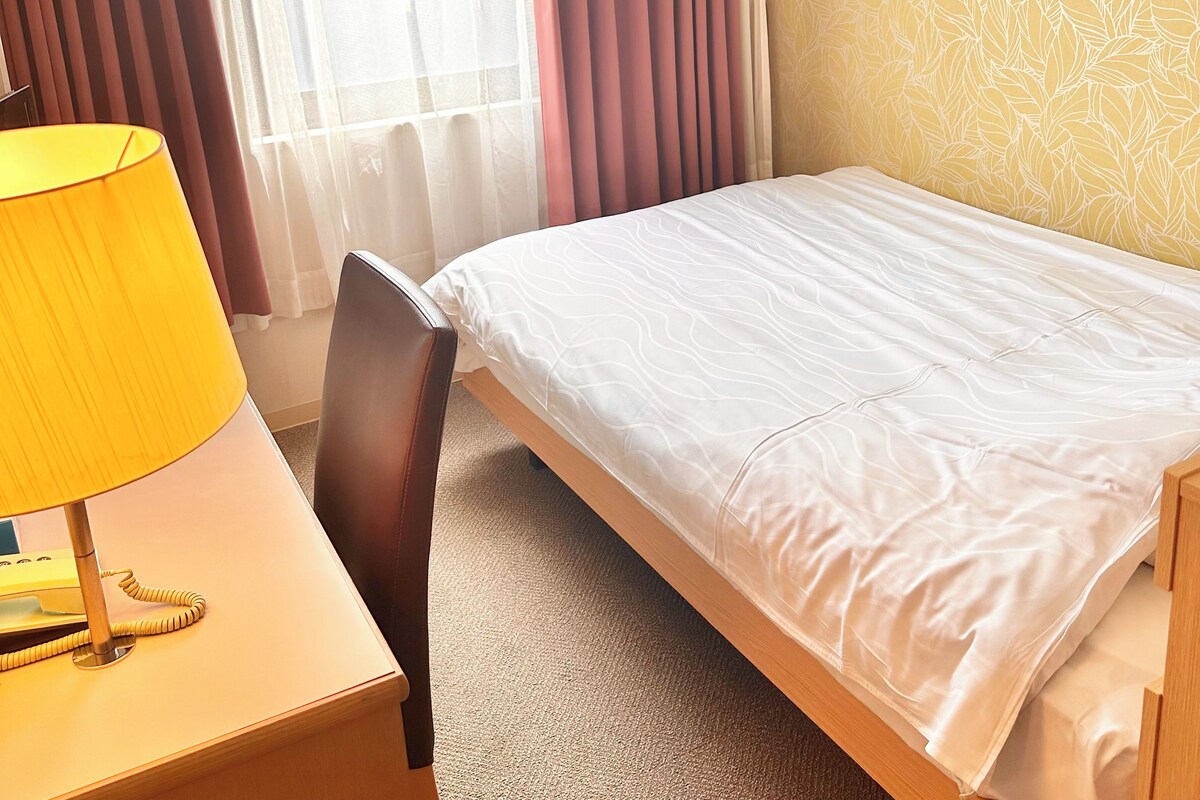 Quarto de hotel com cama, escrivaninha com luminária e uma cadeira.