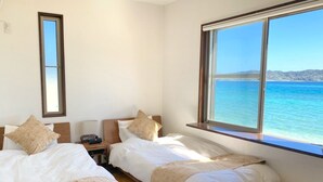 Quarto de hotel com duas camas, mesa de cabeceira, luminária e uma janela grande com vista para o mar.