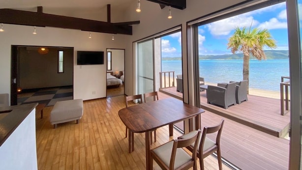 Interior moderno com piso de madeira, área de jantar com mesa e cadeiras, e janelas amplas com vista para o mar e palmeiras.