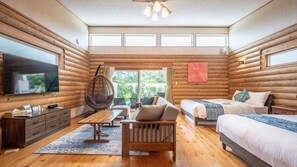 Interior de una cabaña de troncos con televisor, sofá, sillón colgante y cama con vistas al exterior.