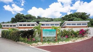 Un letrero de hotel con el nombre 'Tabino Hotel Villa Miyakojima' rodeado de exuberante follaje y un cielo despejado.