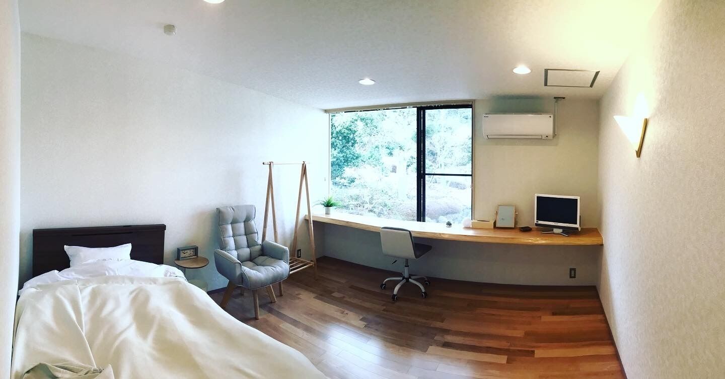A modern bedroom with a bed, a desk with a computer, a chair, and a window with a view of trees.