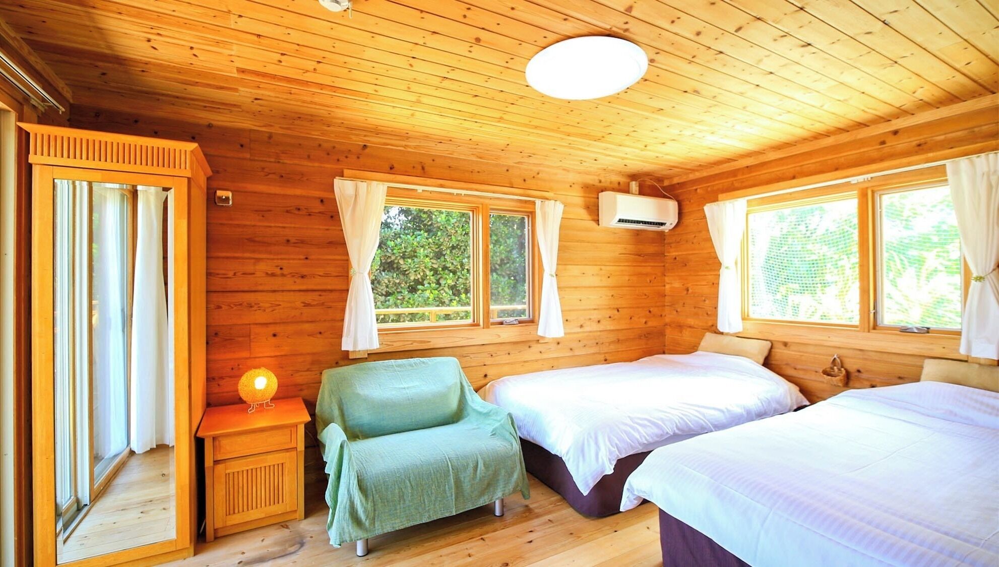 A wooden cabin room with two beds, a chair, a nightstand, and a window with curtains.