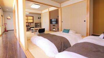 A traditional Japanese-style hotel room with tatami flooring, a television, and a dining area visible through a sliding door.