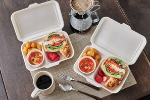 A meal setup with two open food containers, a cup of coffee, and a coffee maker on a wooden table. - The Canvas Hayama Park (Hayama)