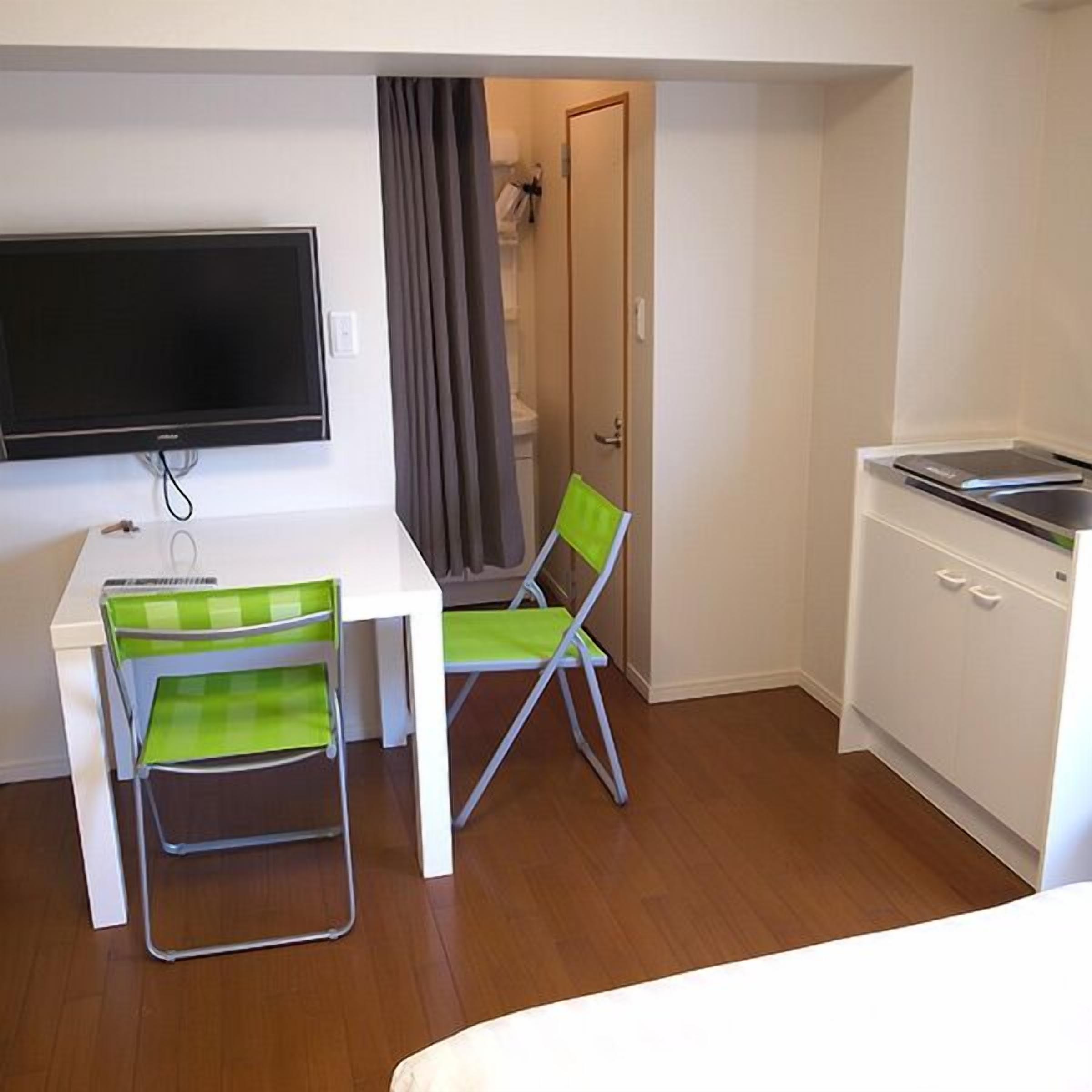A compact room with a white desk, two green chairs, a television, a door, and a small kitchenette.