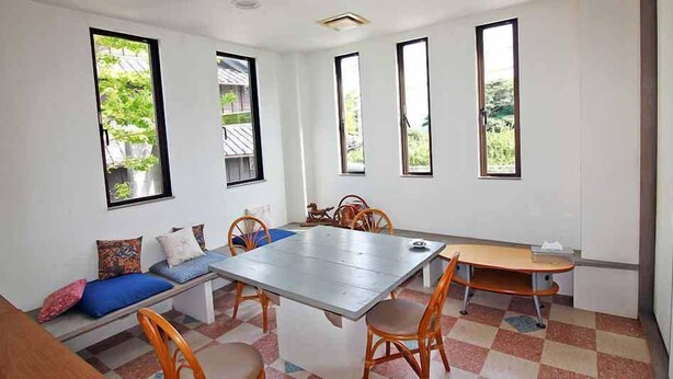 Comedor con una mesa grande, sillas y un banco con cojines. Cuatro ventanas permiten la entrada de luz natural.