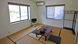 Una habitación con suelo de tatami, una mesa baja con cojín, un televisor sobre un mueble y una ventana con vistas a edificios.
