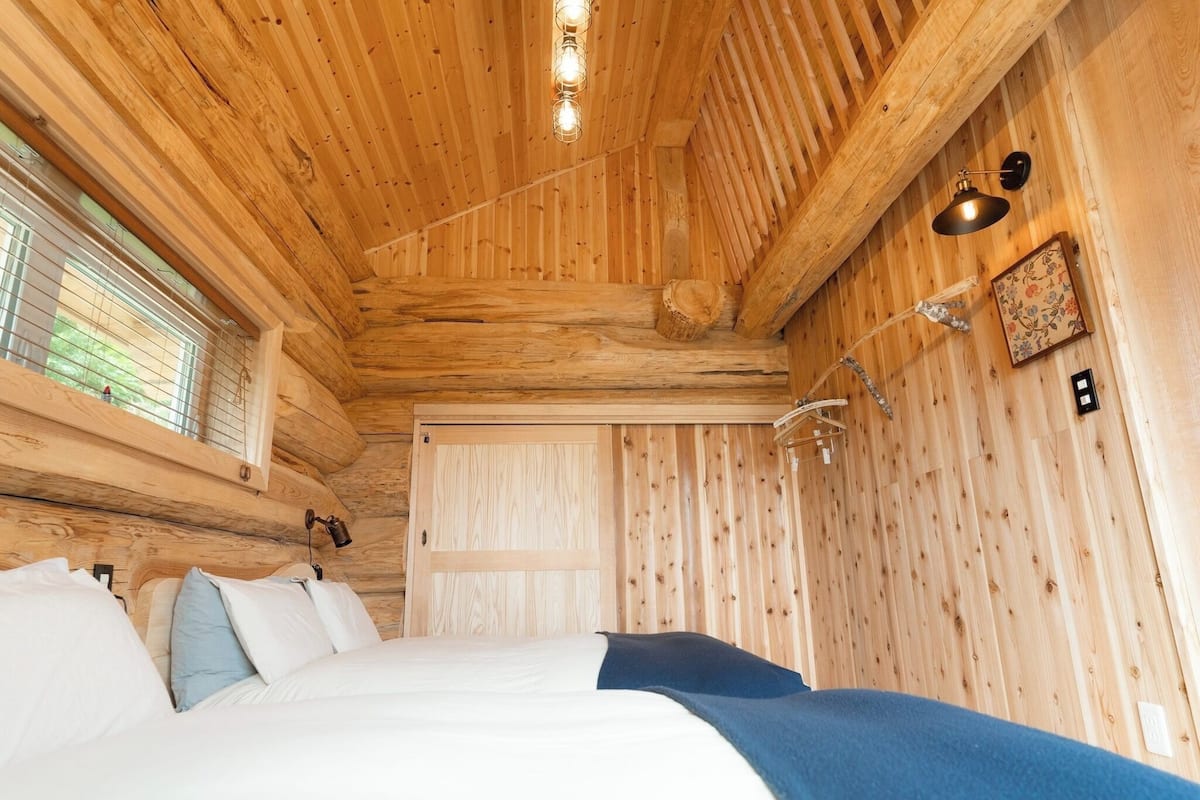 A wooden cabin bedroom with a bed, a window with blinds, and a wall-mounted light fixture.