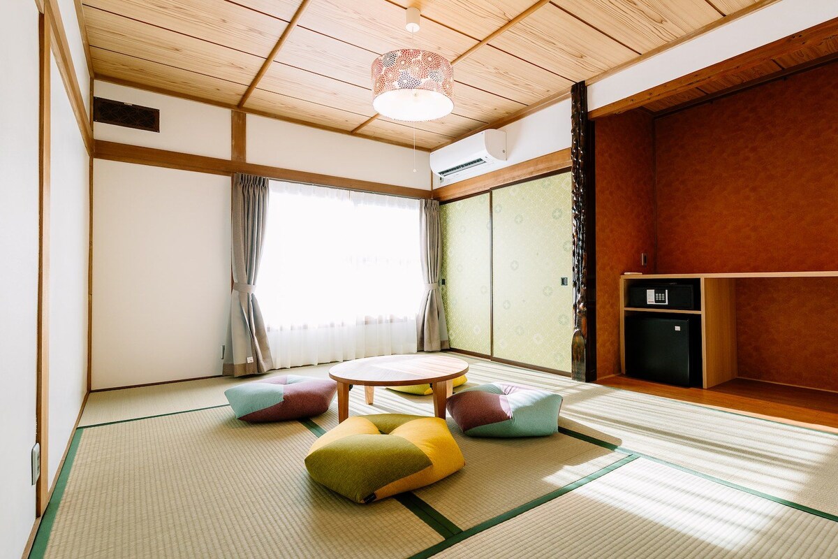 Une pièce de style japonais traditionnel, avec un sol recouvert de tatamis, une petite table basse, des coussins, un petit réfrigérateur et une porte coulissante.