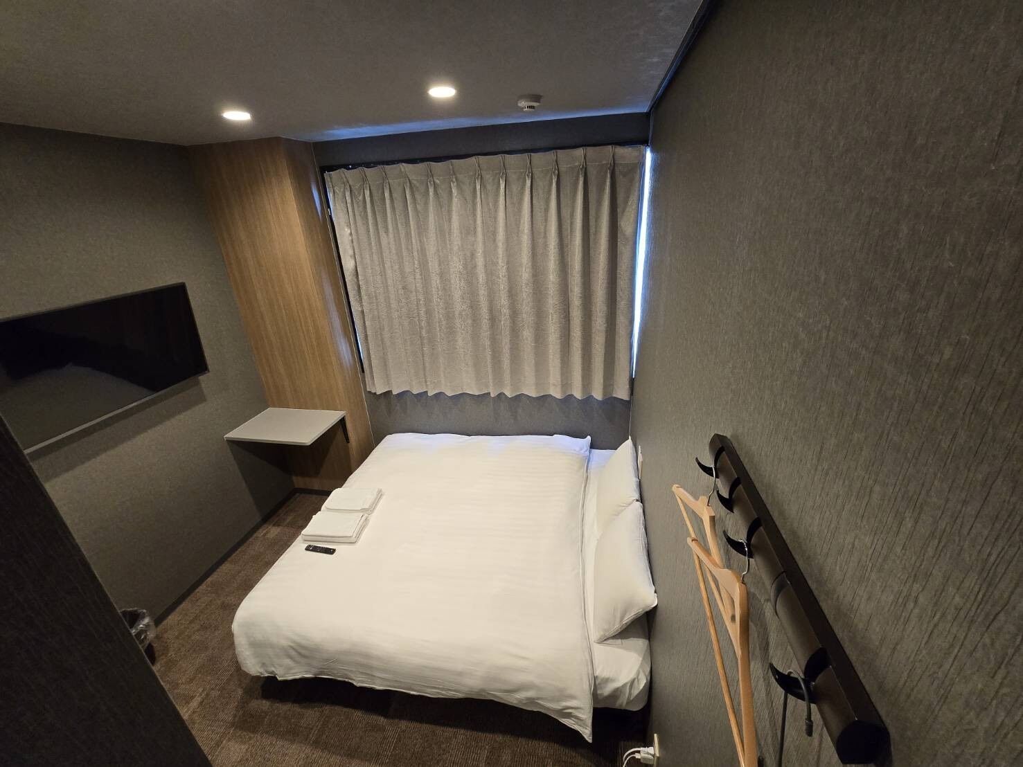 A hotel room with a bed, a bedside table, a TV mounted on the wall, and a window with curtains.