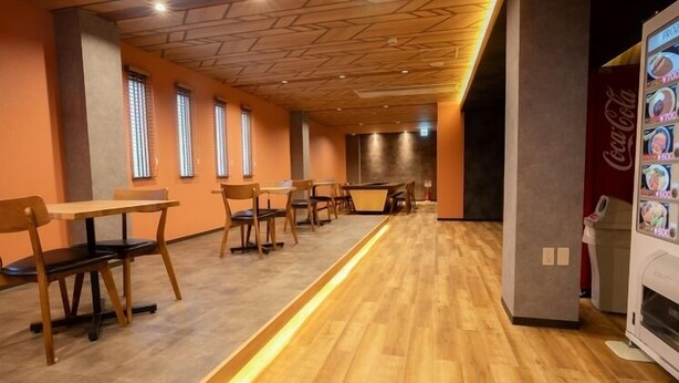 A modern dining area with wooden tables and chairs, a Coca-Cola vending machine, and a long wooden bench along the wall. - Ping Pong Hotel & Cabin Tokuyama Ekimae / Ping Pong Residence Kojimachi (Shunan)