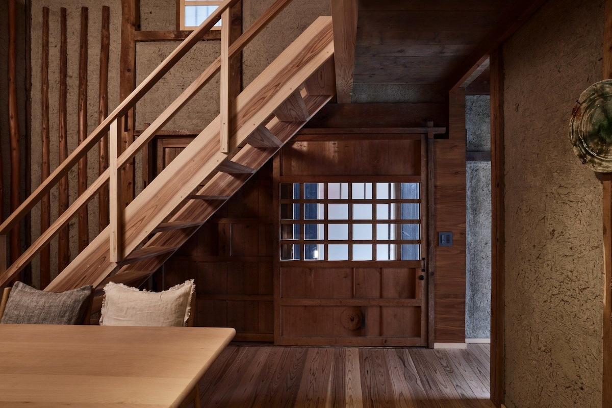 Una escalera de madera que lleva a una puerta con diseño de rejilla, una mesa con cojín y una pared rústica.