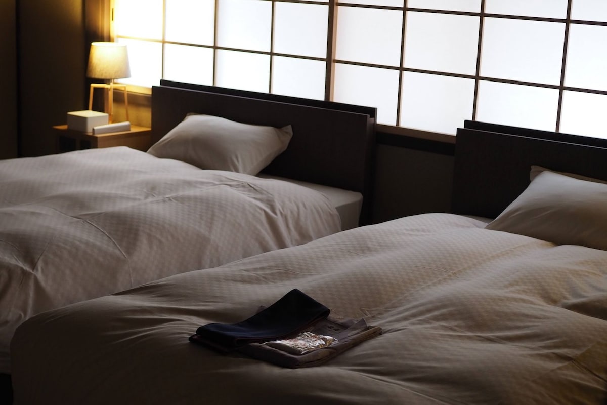 Habitación de hotel con dos camas, una mesita de noche con lámpara y una ventana con diseño de cuadrícula.