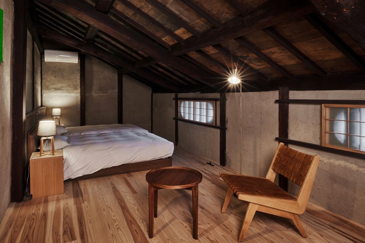 A rustic room with a wooden ceiling, a bed, a chair, a nightstand, a lamp, and a window with a view.