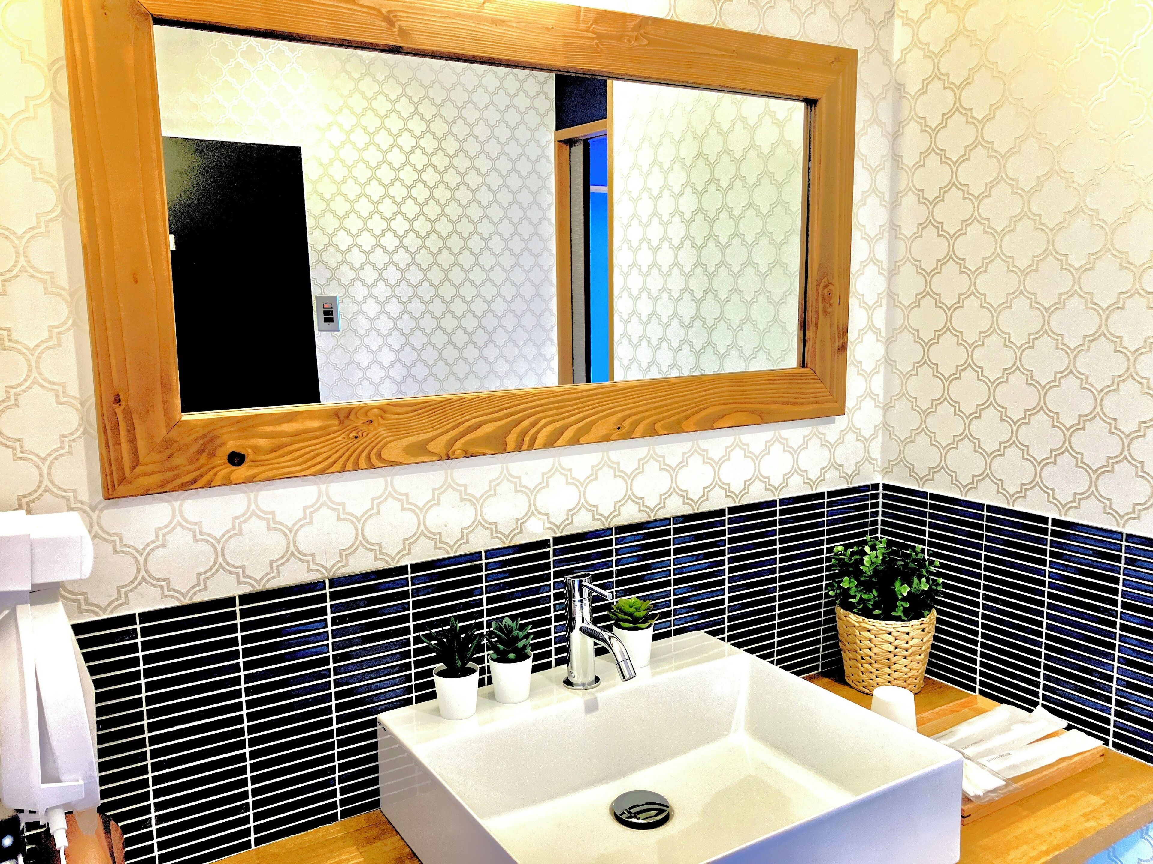 Ein Badezimmer mit einem weißen Waschbecken, zwei Blumentöpfen, einem Spiegel mit Holzrahmen und einer Wand mit Muster.