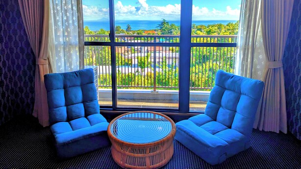 Une pièce avec deux fauteuils bleus, une table basse en osier et une grande fenêtre donnant sur un balcon et la mer.