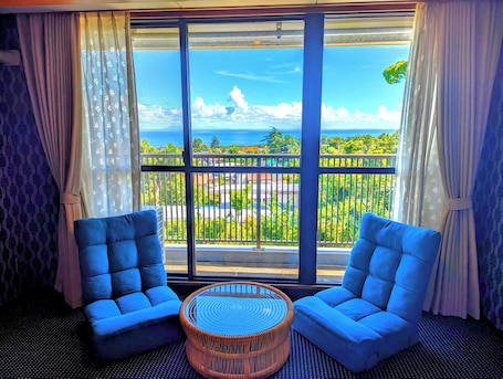 A room with two blue armchairs, a wicker coffee table, and a large window with a view of a balcony and the sea.