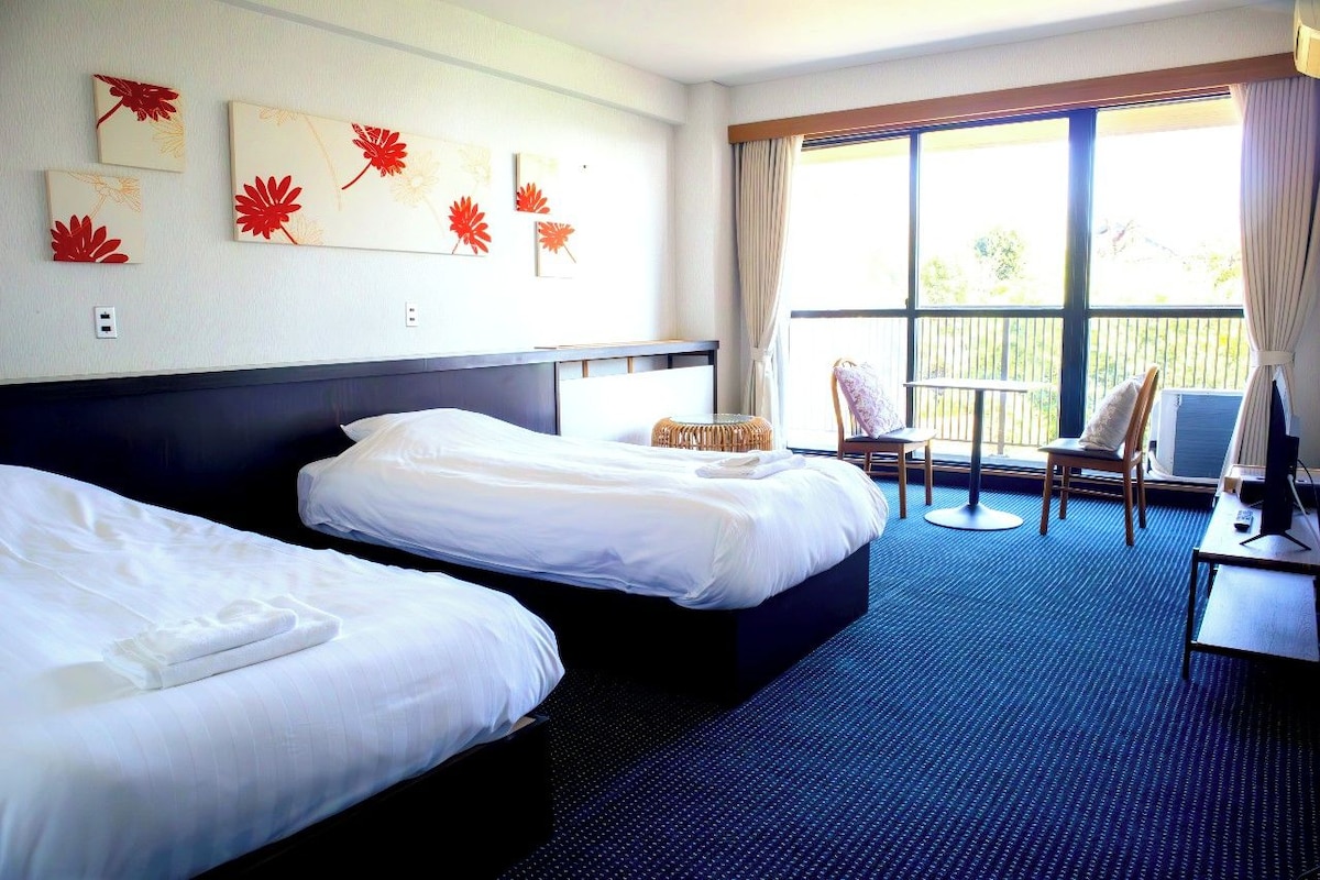 A hotel room with two beds, a small table with a chair, and a balcony with a view.