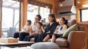 A family sitting on a leather couch in a well-lit room with large windows, a wooden ceiling, and a table with a book and a cup. - Mori no Kiki (Minamioguni)