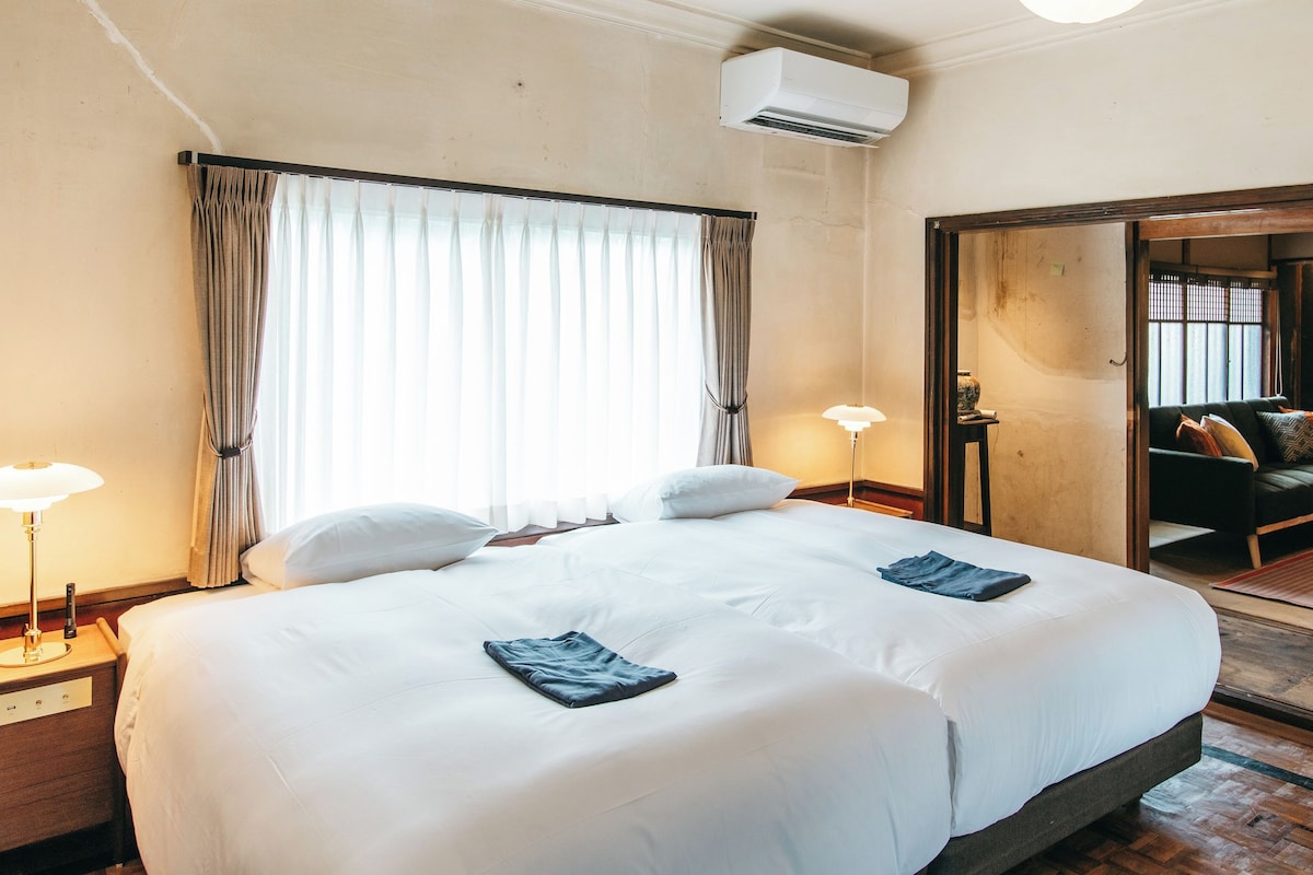 A hotel room with a large bed, bedside lamps, an air conditioning unit, and a view into a living area with a sofa and cushions.