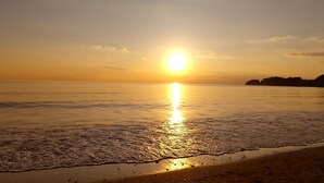 Una playa con el sol reflejado en el agua, olas mansas lamiendo la orilla y una masa de tierra lejana silueteada contra el cielo.