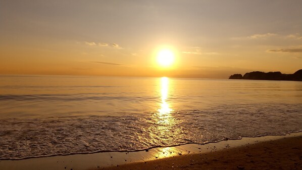 A beach with the sun reflecting on the water, waves gently lapping at the shore, and a distant landmass silhouetted against the sky. - Kohaku Amber Kamakura Zaimokuza (Kamakura)